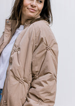 Model wearing a taupe puffer jacket with flower detail, collared neckline and long sleeves.