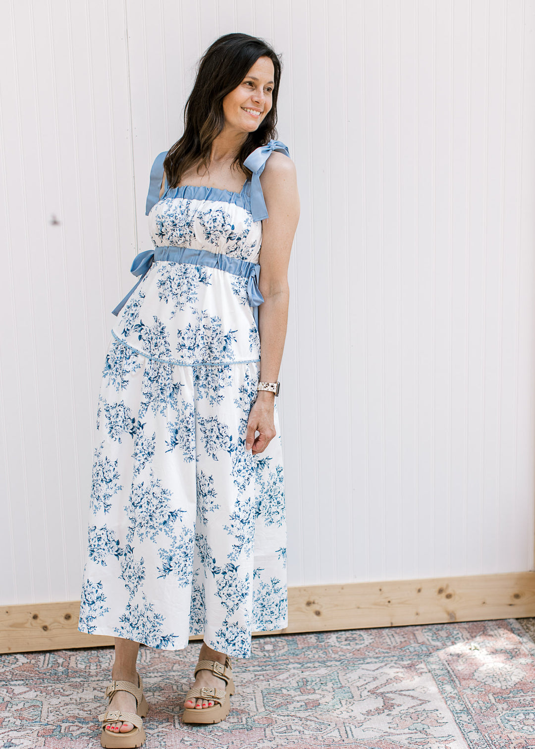 Model wearing wedges and a white midi with blue floral, elastic at bust and tie spaghetti strap.
