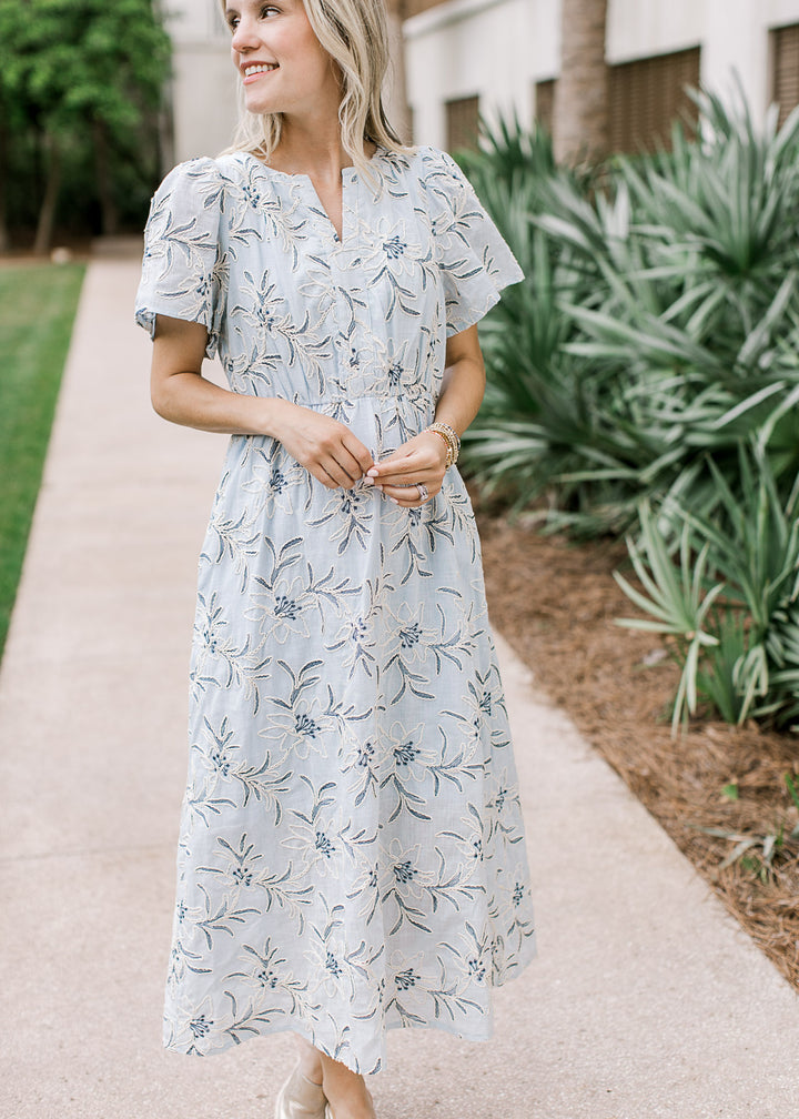 Model wearing a pale blue midi with cream and navy floral,  v-neck, short sleeves and pockets.