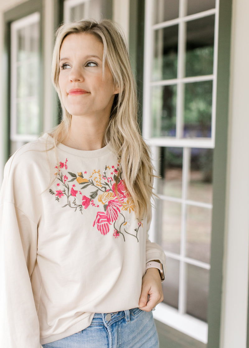 Model wearing a cream top with embroidered flowers, long sleeves and a round neck. 