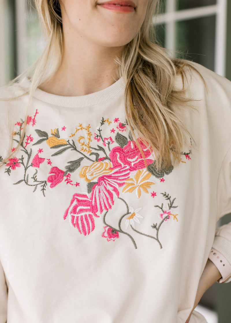 Close up of pink, yellow and green embroidered flowers on a cream top with long sleeves.