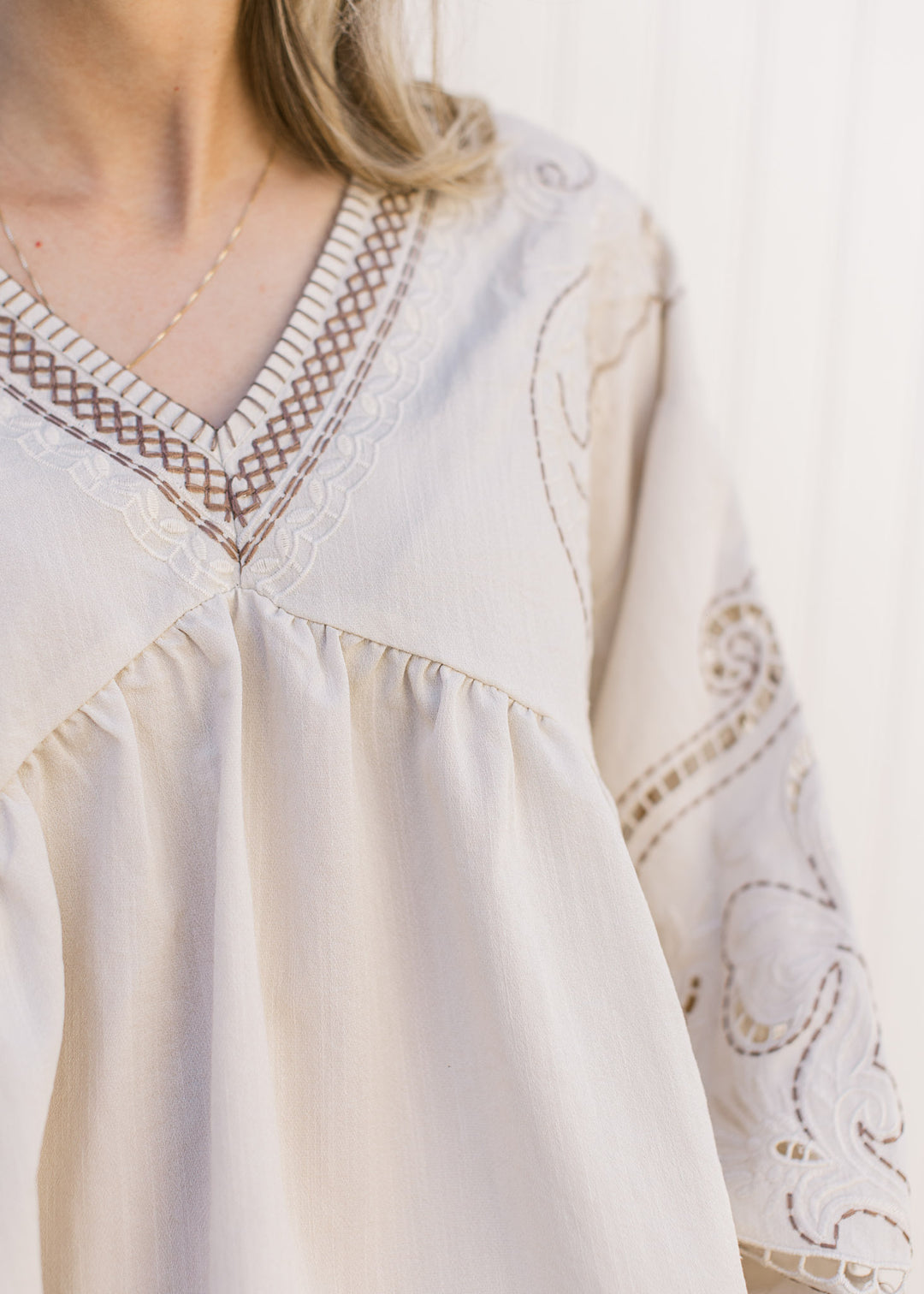 Close up of taupe and cream embroidery on the v-neck of a khaki colored top with long sleeves. 