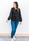 Model wearing jeans, sneakers and a black long sleeve top with white embroidered flowers. 