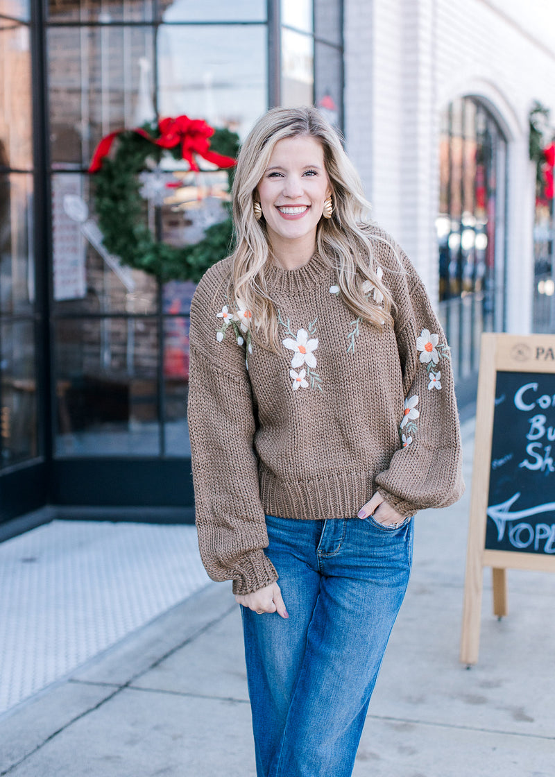 Model wearing jeans and an olive green sweater with cream embroidered flowers and long sleeves. 