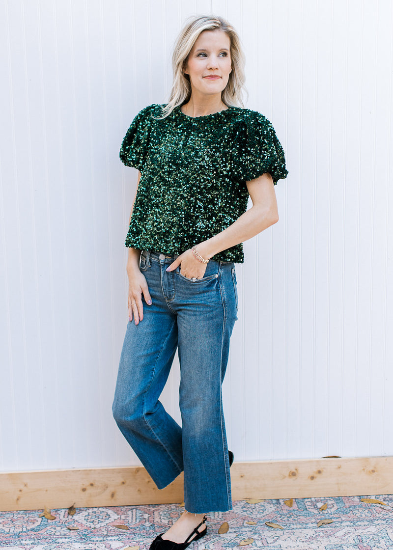 Model wearing jeans with an emerald green sequin top with short puff sleeves and pleated shoulders.