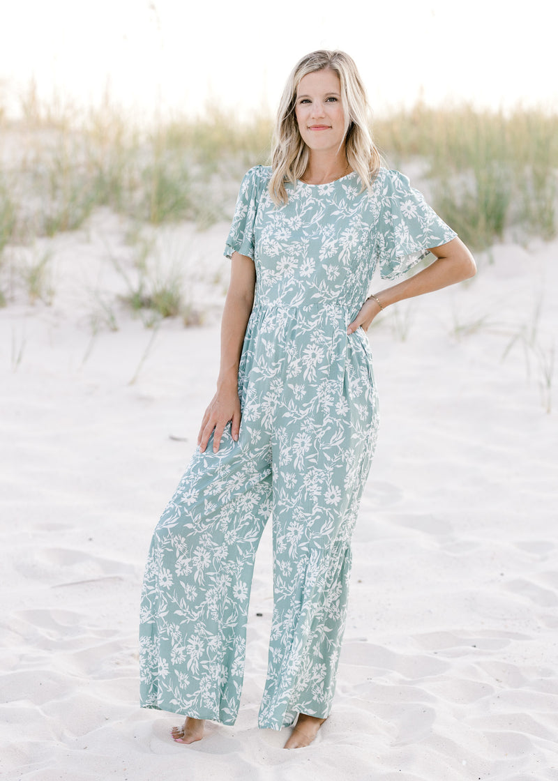 Model wearing a wide leg sage jumpsuit with white floral pattern and short sleeves. 
