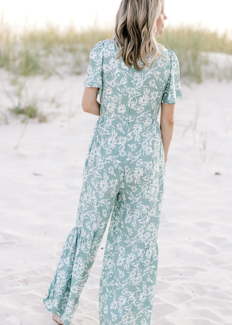 Back view of model wearing a sage jumpsuit with white floral pattern, short sleeves and zip closure.