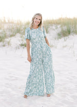 Model wearing a wide leg sage jumpsuit with white floral pattern with a smocked detail at the waist.