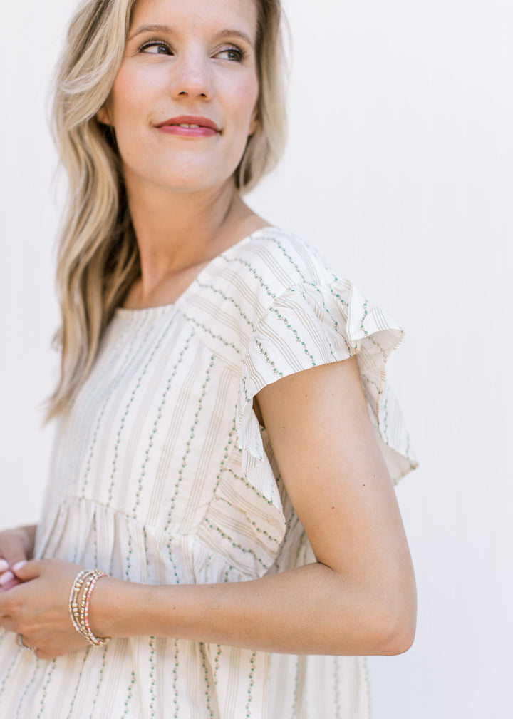 Model wearing a cream top with tan and green ivy pin stripes, a square neck and ruffle cap sleeve. 