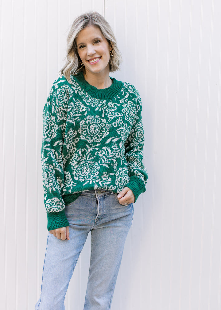 Model wearing jeans and an evergreen sweater with off-white floral, long sleeves and evergreen trim.