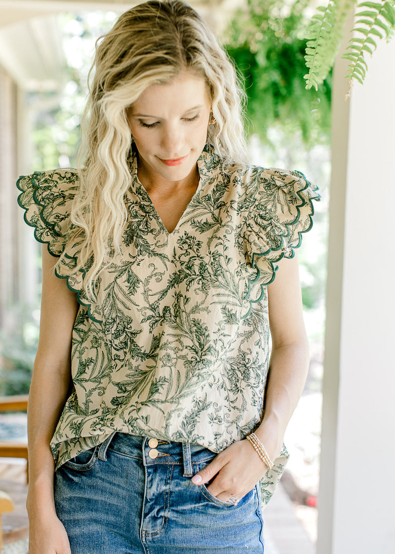 Model wearing a beige top with green floral, ruffle cap sleeves and a v-neck. 