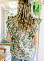 Back view of Model wearing a beige top with green floral, ruffle cap sleeves and a v-neck. 