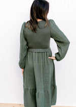 Back view of Model wearing an evergreen dress with bubble long sleeves and ruffle at waist. 