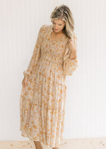 Model wearing a taupe maxi dress with a light floral, smocked bodice and sheer long sleeves. 