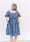 Back view of Model wearing a medium washed denim dress with bubble short sleeves and a square neck.