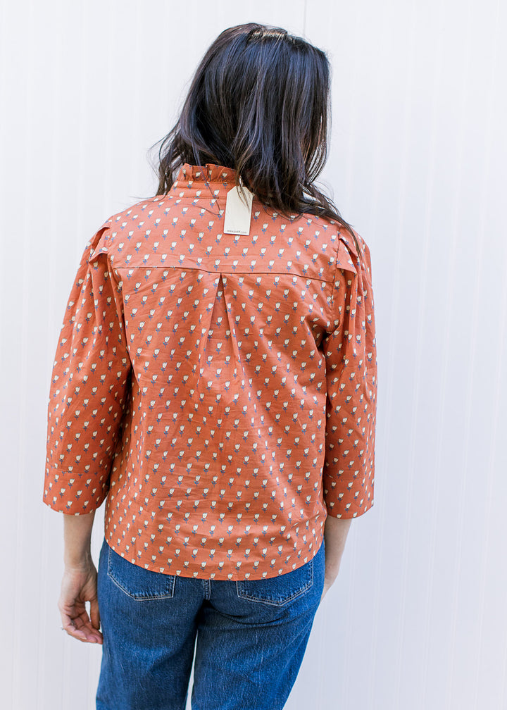 Back view of model wearing a rust top with soft yellow floral print ruffle at neck and 3/4 sleeve. 