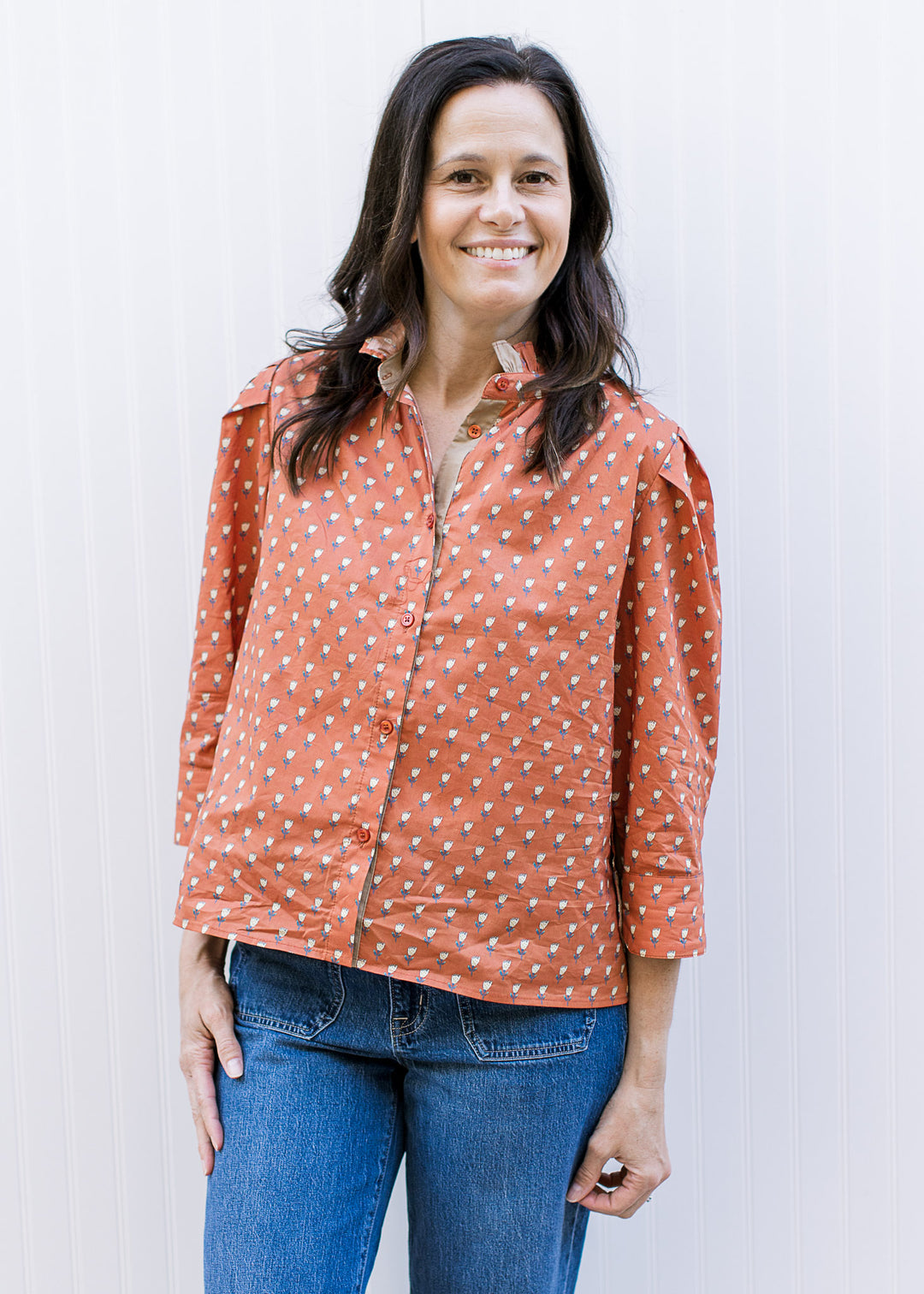 Model wearing jeans and a rust top with soft yellow floral, ruffle at neck and 3/4 sleeve.