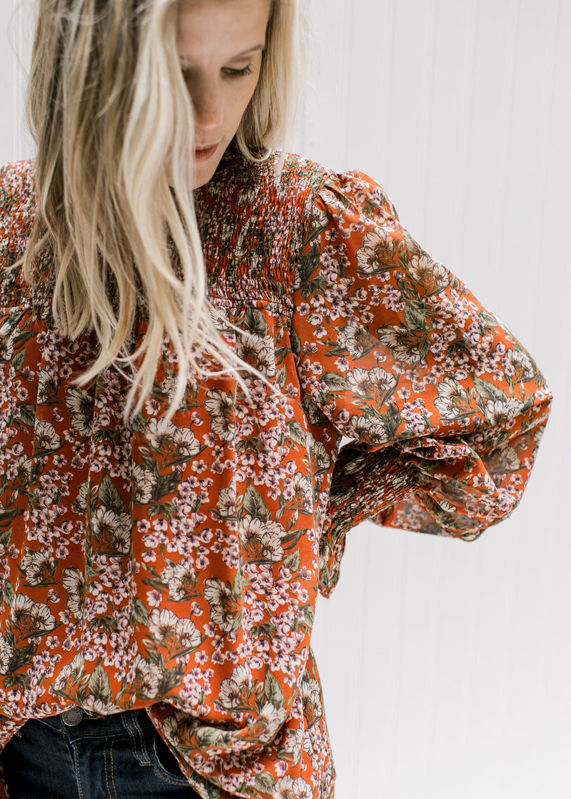 Model wearing a fully lined rust top with a cream floral, sheer long sleeves and a mock neck. 