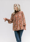 Model wearing jeans with a rust top with a cream floral, sheer long sleeves and a mock neck. 