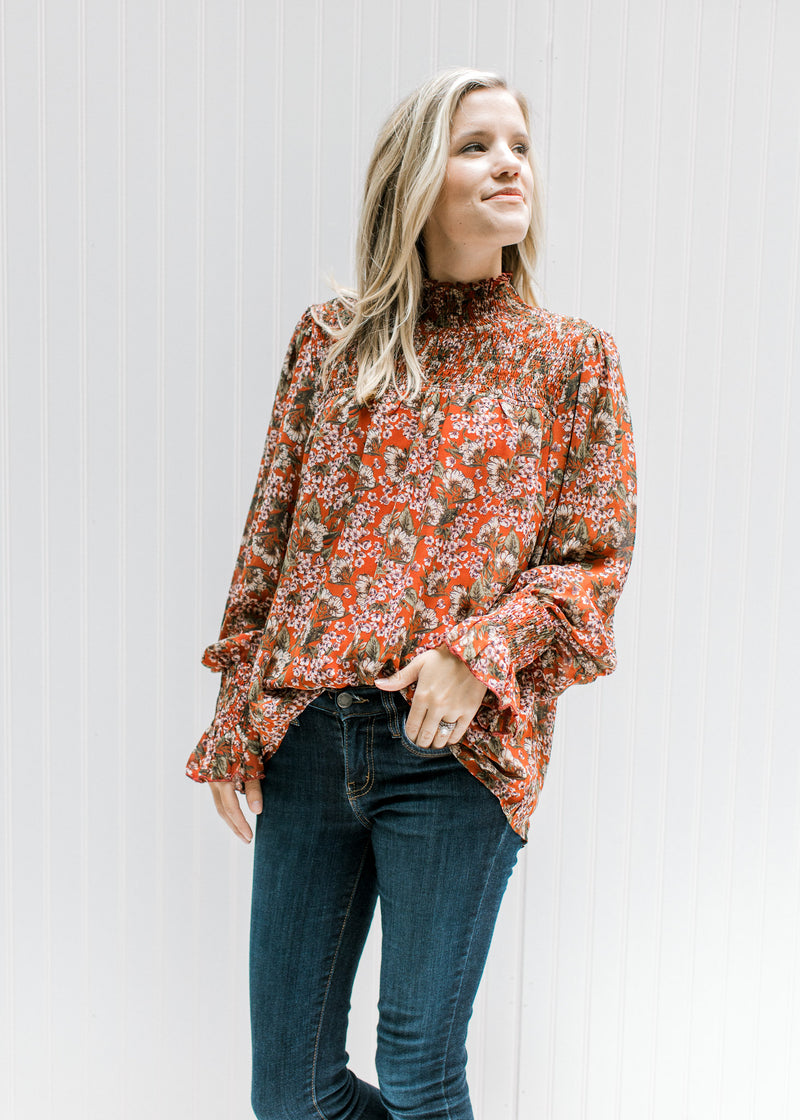 Model wearing a rust top with a cream floral, sheer long sleeves and a mock neck with smocked yolk. 