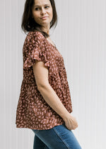 Model wearing a rust top with a pink floral pattern and an extended shoulder with a ruffle. 