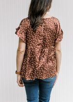 Back view of model wearing a rust top with light pink floral pattern and short sleeves with ruffle.