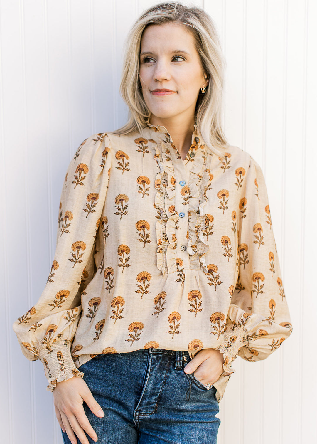 Model wearing a camel top adorned with mustard floral, button placket with ruffle and long sleeves.