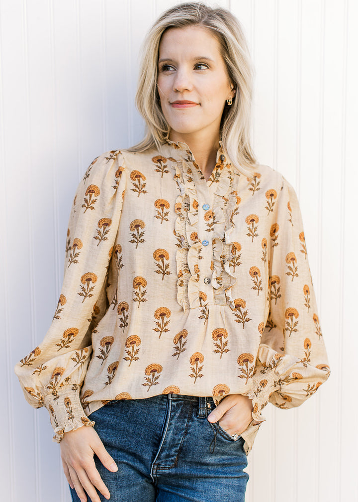 Model wearing a camel top adorned with mustard floral, button placket with ruffle and long sleeves.