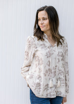 Model wearing a cream top with taupe flowers, v-neck with a tie and long sleeves with button cuff.