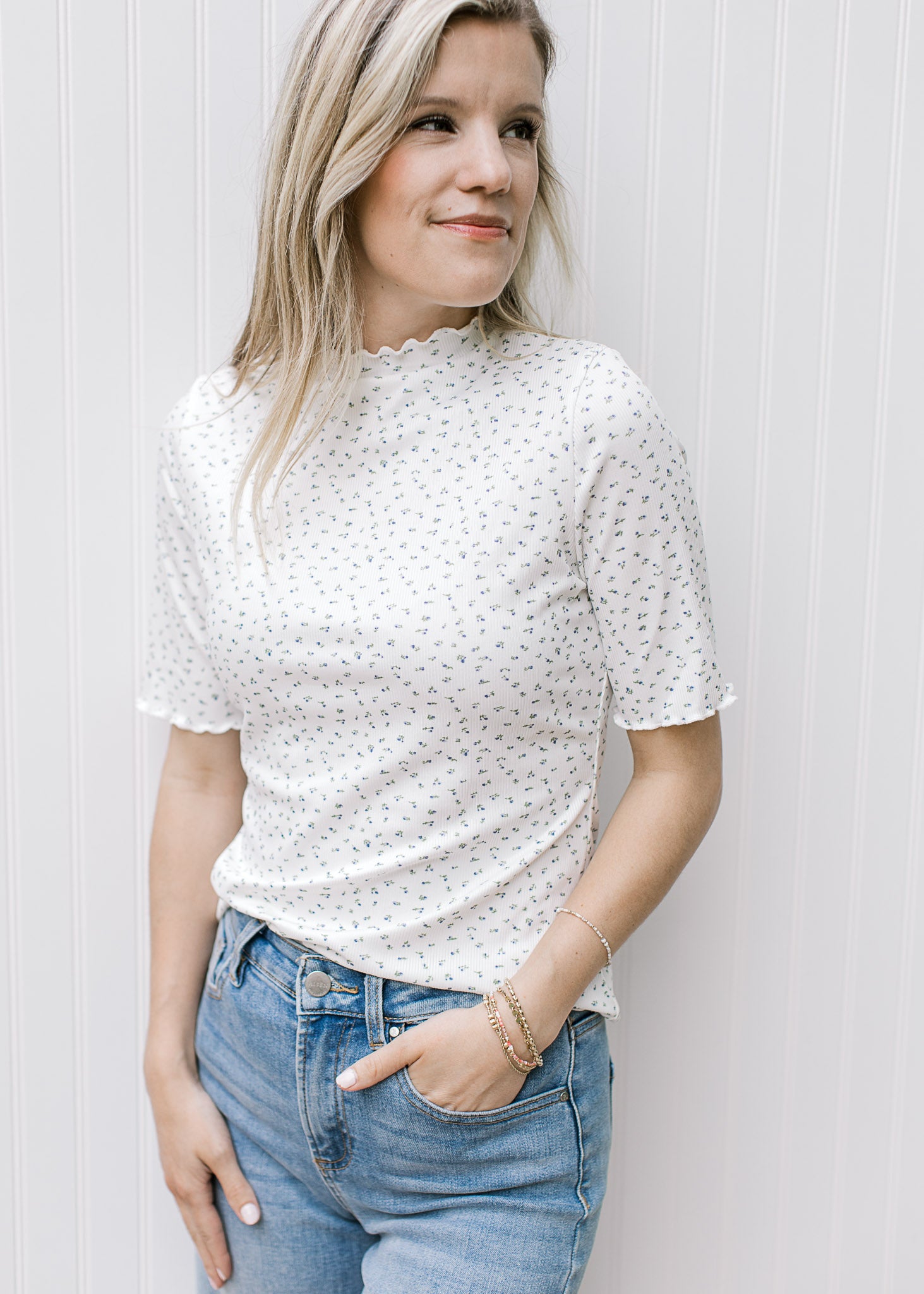 Model wearing a ribbed cream floral top with a ruffle at short sleeve, hem and mock neck.