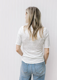 Back view of Model wearing a cream floral top with a ruffle at short sleeve, hem and mock neck.