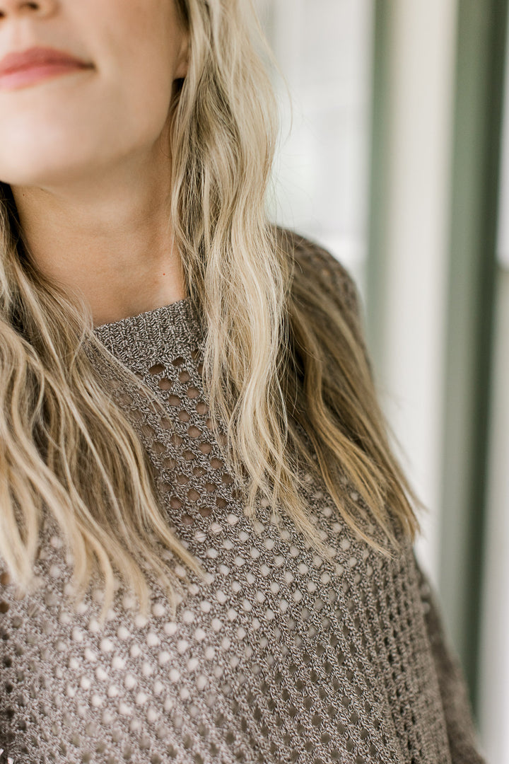 Close up of loose knit material on a brown sweater with a round neck and long sleeves.