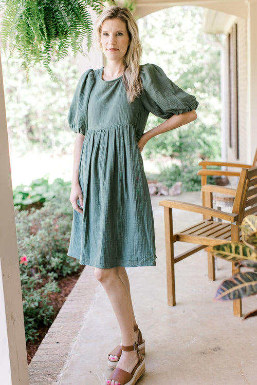 Model wearing heels with a deep sage, gauze dress hitting above the knees with bubble short sleeves.