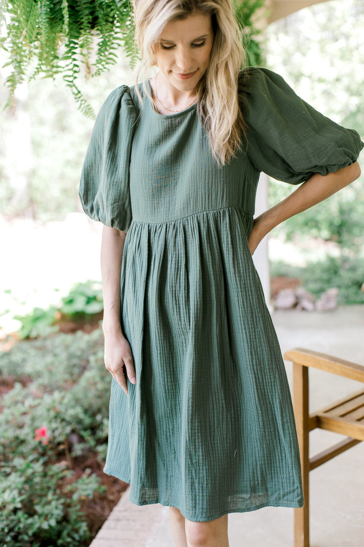 Model wearing a deep sage, gauze, knee length dress with bubble short sleeves.