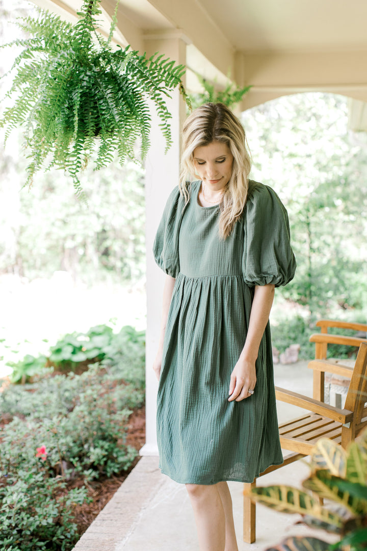 Model wearing a deep sage, gauze dress hitting just above the knees with short sleeves.