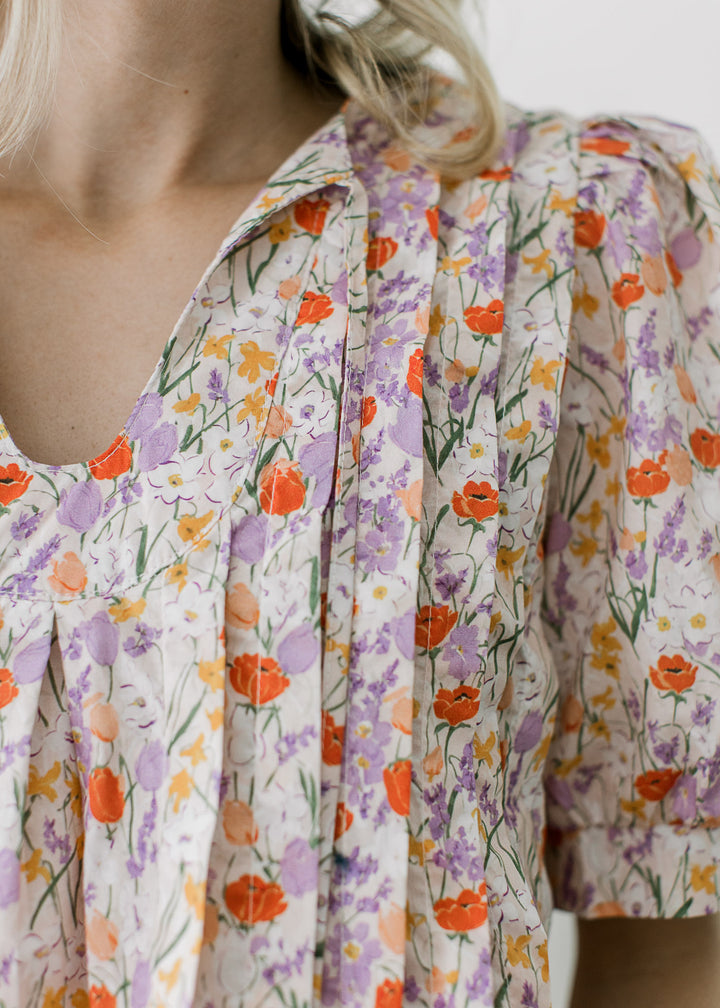 Close up on pleating and v-neck with tie on off-white top with purple, rust, and yellow floral.