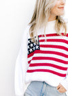 Model wearing a white lightweight knit sweater with a flag across the front and long sleeves.