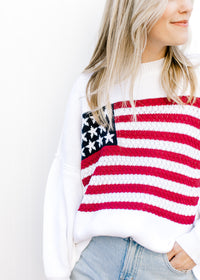Model wearing a white lightweight knit sweater with a flag across the front and long sleeves.