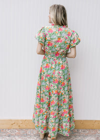 Back view of Model wearing a bright floral maxi with short sleeves, elastic waist and ruffle neck.