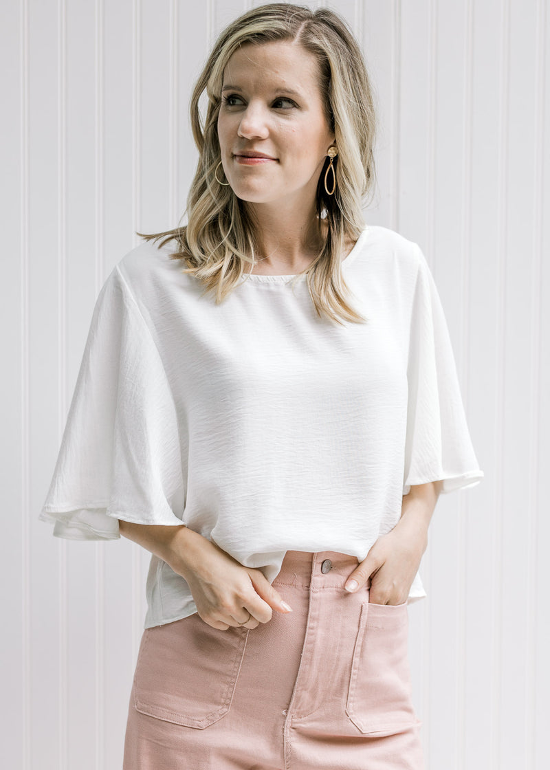 Model wearing pink pants with an ivory polyester slightly cropped top with flutter short sleeves. 