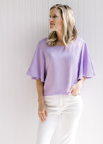 Model wearing white jeans with a light lavender slightly cropped top with flutter short sleeves. 