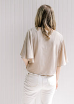 Back view of model wearing a polyester taupe crop top with flutter short sleeves and a flowy fit. 