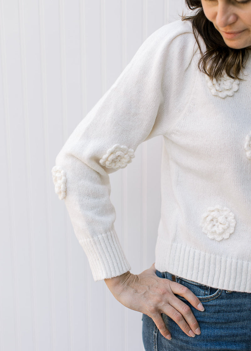 Model wearing a cream knit sweater with appliqué flowers, long sleeves and pleated shoulders. 