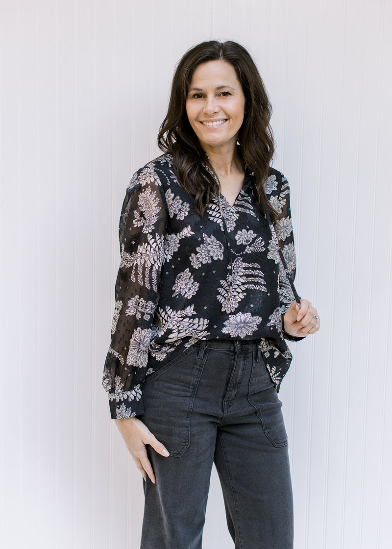 Model wearing a black top with a gray floral pattern, sheer long sleeves and v-neck with a tie. 