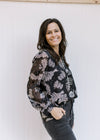Model wearing a black top with gray floral, sheer long sleeves and a v-neck with a ruffle and a tie.