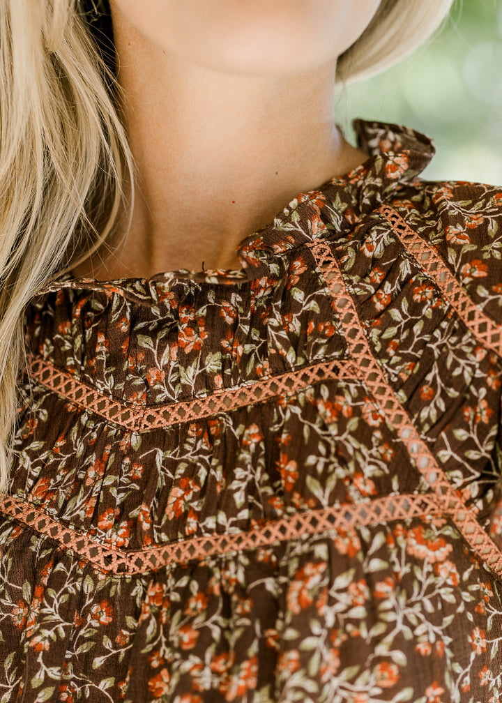 Close up view of ruffle neck and lace at bodice of a brown top with maroon and green floral. 