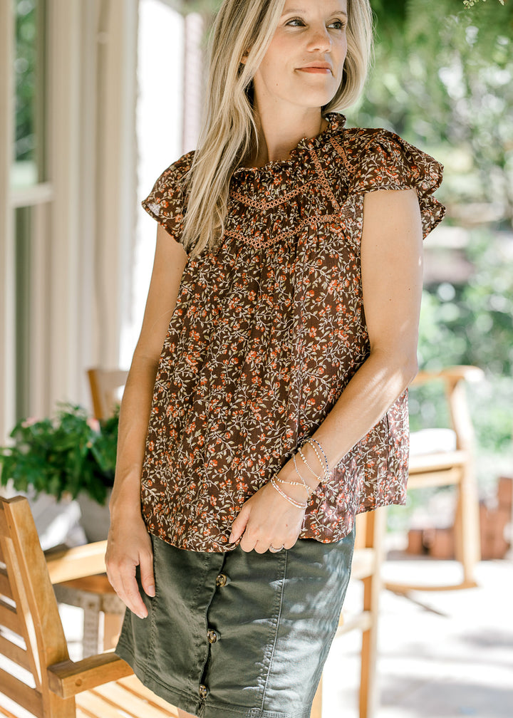 Model wearing a brown top with maroon and green floral, flutter sleeves, and lace detail at bust.