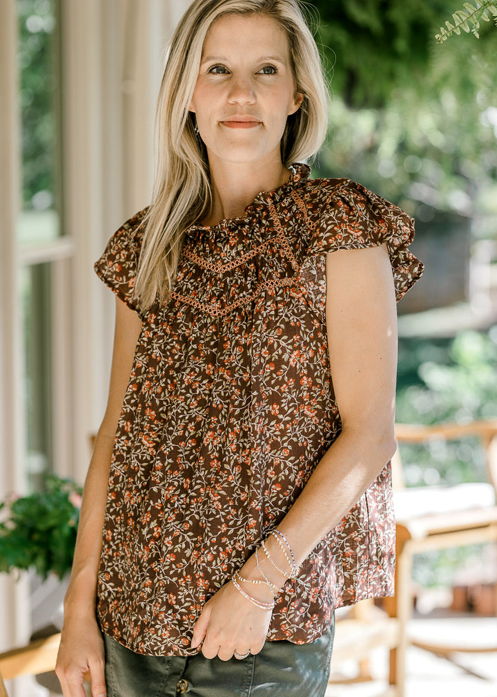 Model wearing a brown top with maroon and green floral, flutter cap sleeves, and ruffle at neck.
