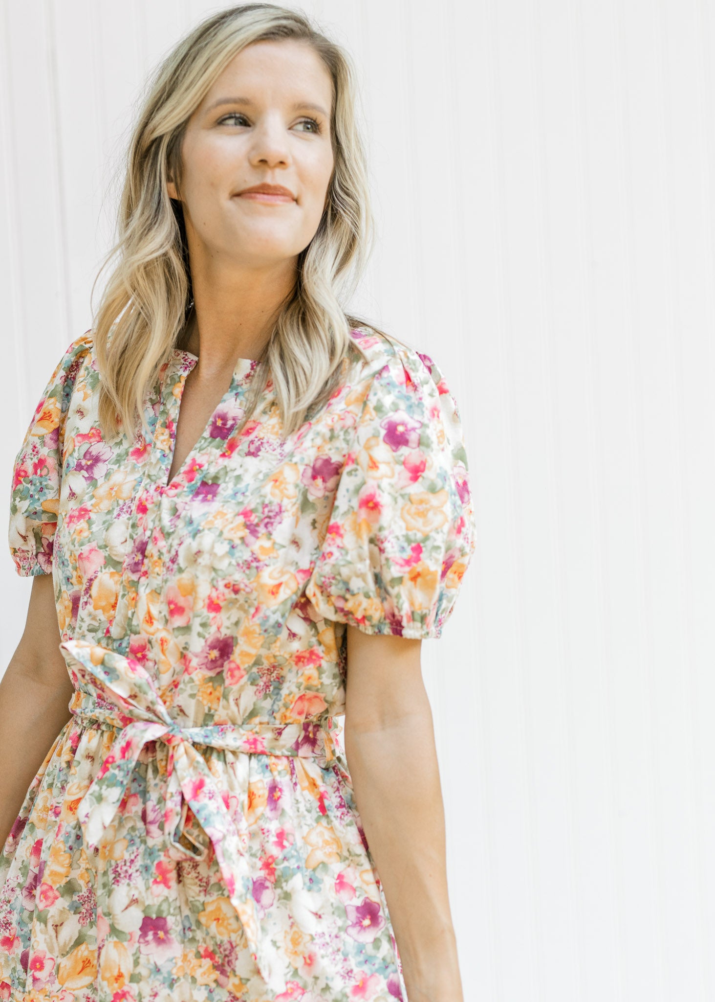 Model wearing a cream v-neck dress with a mixed colored floral pattern and a tie at the waist. 