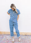Model wearing a medium washed denim jumpsuit with a hidden zipper, wide legs and short sleeves. 